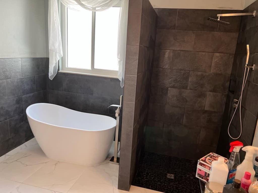 Bathroom freestanding soaking tub left glass partition charcoal tile shower rainfall head pebble floor chrome fixtures daylight.