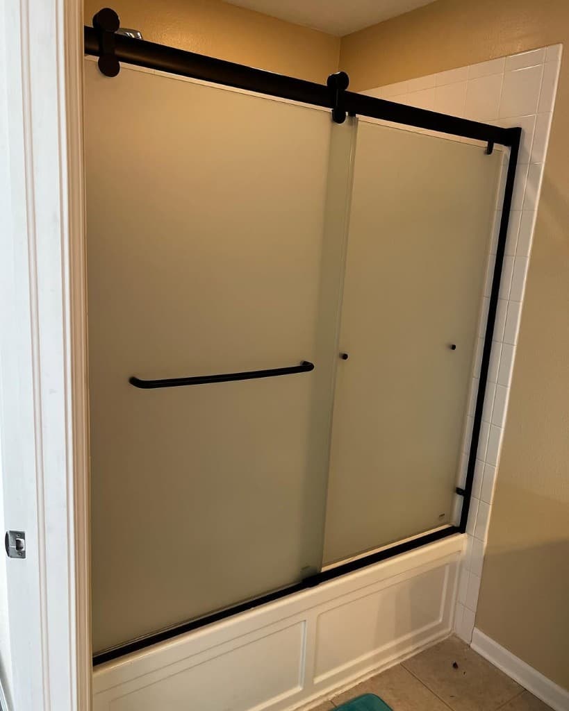 Sliding frosted shower doors matte black fittings over white apron tub with subway tile backdrop.