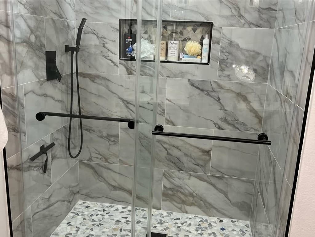 Walk shower grey veined porcelain walls black fixtures pebble mosaic shower floor niche toiletries glass enclosure.