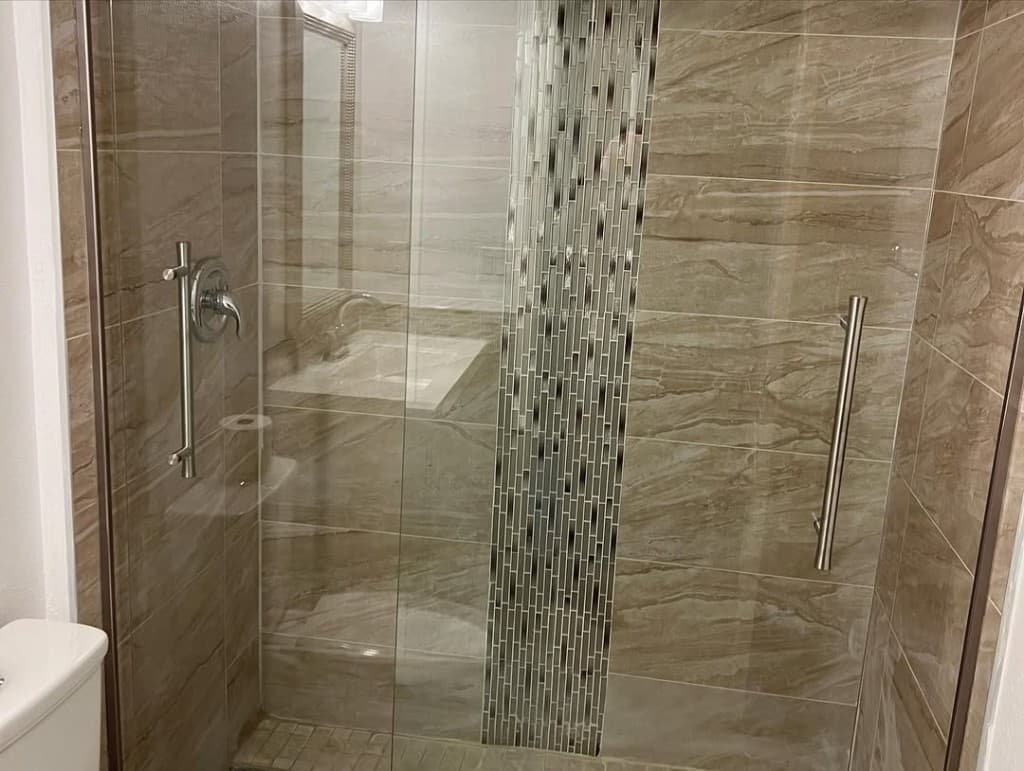 Walk shower glass door marble walls vertical mosaic brushed metal handles mosaic floor vanity visible through doorway.