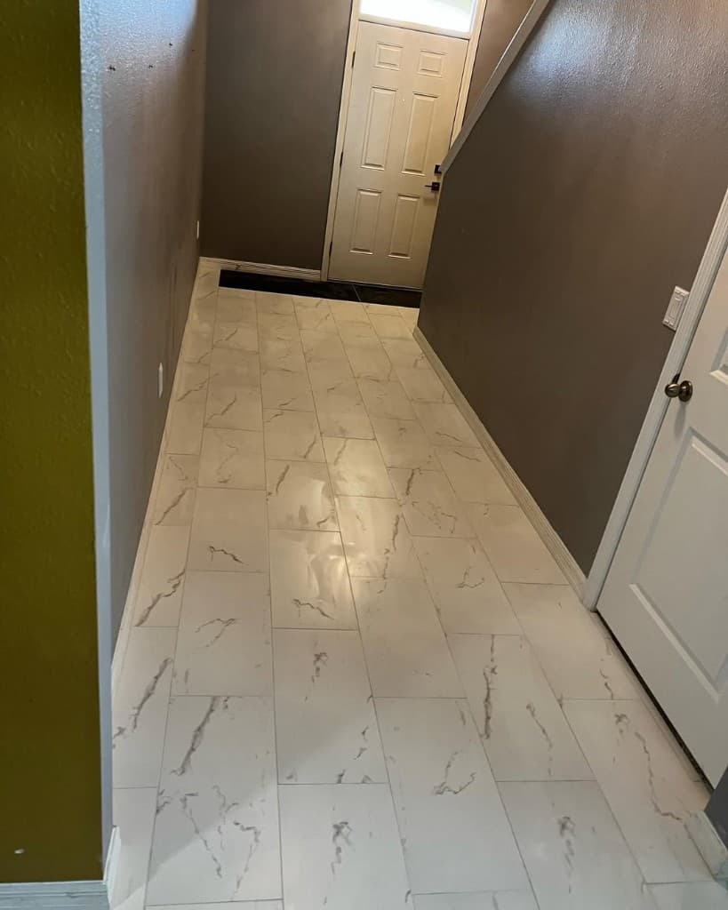 Long residential hallway glossy white veined porcelain tile staggered lay leading to paneled interior door sunny.