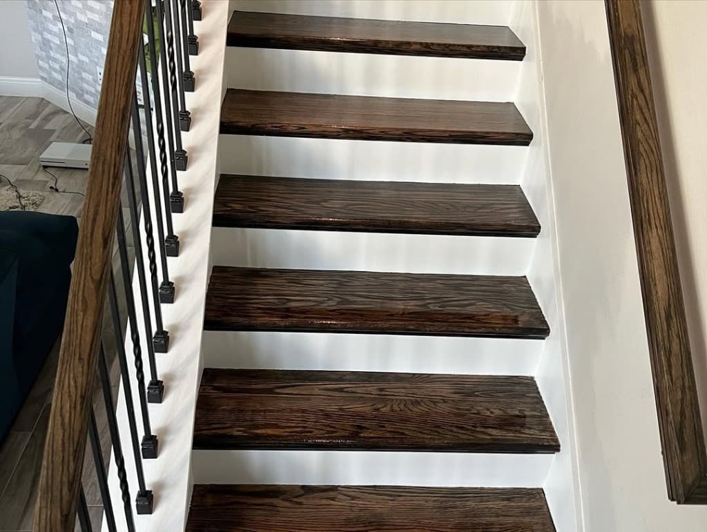 Looking down staircase dark glossy wood tread white risers black twisted metal balusters handrails grey floor living below.