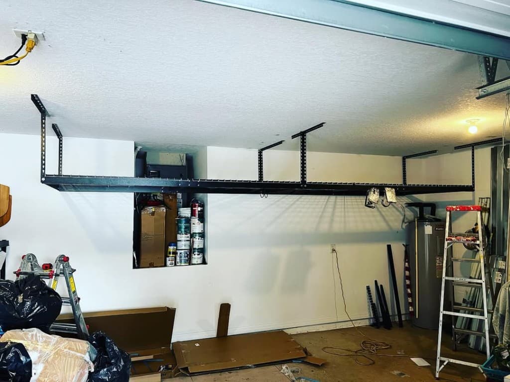 Wide garage overhead black rack ladders paint supplies cord concrete floor cluttered active jobsite fluorescent bulb.