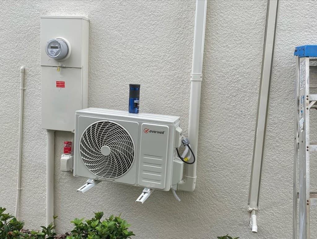 White Everwell outdoor mini-split meter disconnect conduit ladder shrubs stucco siding blue sky condenser mounts.
