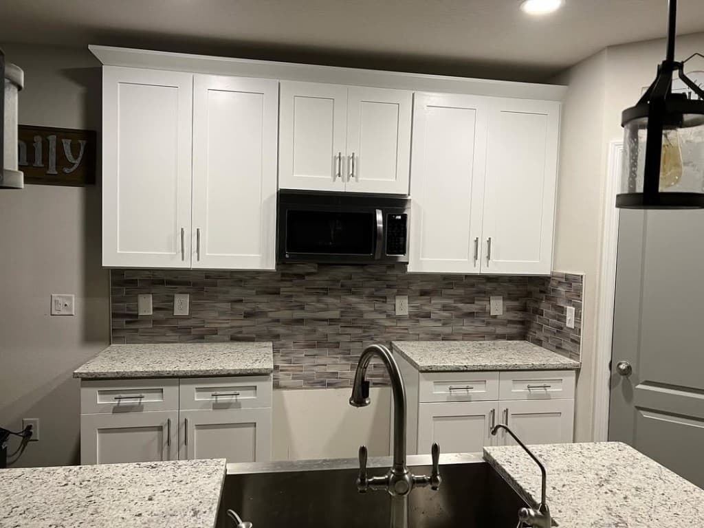 Over island perspective stainless faucet speckled granite mosaic tile wall black microwave shaker white cabinetry crown.