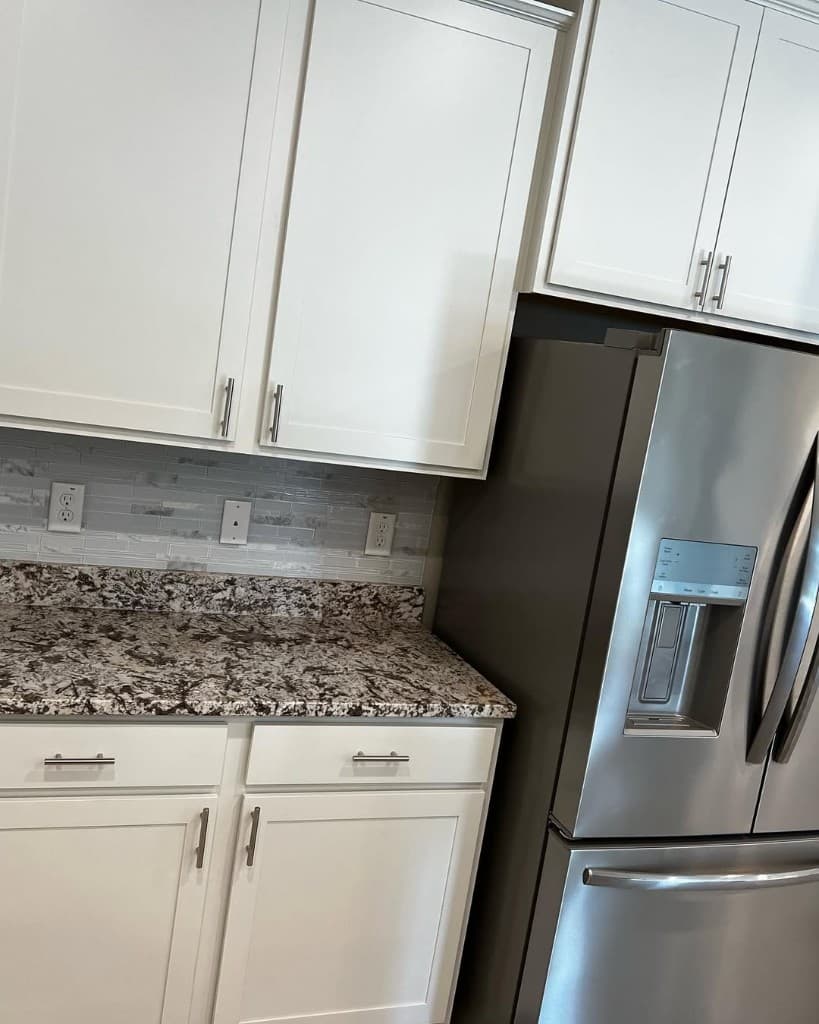 Kitchen corner with white shaker cabinets, grey mosaic backsplash, speckled granite counter, and large stainless fridge.